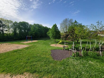Das Bild zeigt den Spielplatz am Bouleplatz in Weilerswist. Das Gras und die Bäume sind grün. Dazwischen stehen Spielgeräte auf Sandflächen. Zu sehen ist eine Schaukel und eine Rutsche. Links mündet ein Weg auf den Spielplatz, der mit Bäumen und Johannisbeersträuchern gesäumt ist.