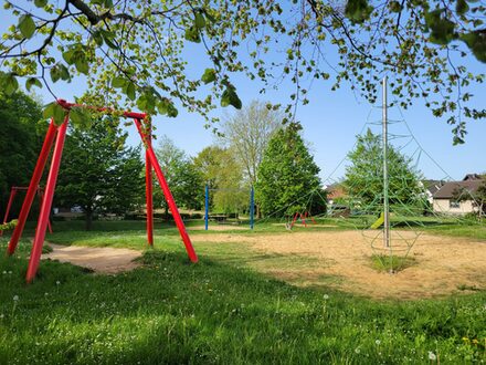 Das Bild zeigt den Spielplatz am Bouleplatz in Weilerswist. Das Gras und die Bäume sind grün. Dazwischen stehen Spielgeräte auf Sandflächen. Zu sehen ist eine Kletterpyramide, eine Seilrutsche und im Hintergrund Schaukel und Rutsche.